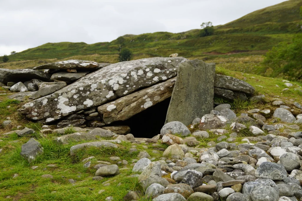 Schwere Steine, die aufeinandergetürmt ein Hügelgrab bilden. Der Eingang führt in die Tiefe des Grabs. im Hintergrund ist eine hügelige Graslandschaft